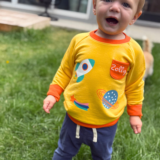 Bright yellow toddler sweatshirt with rocket and planet graphics, featuring an orange collar and pocket.