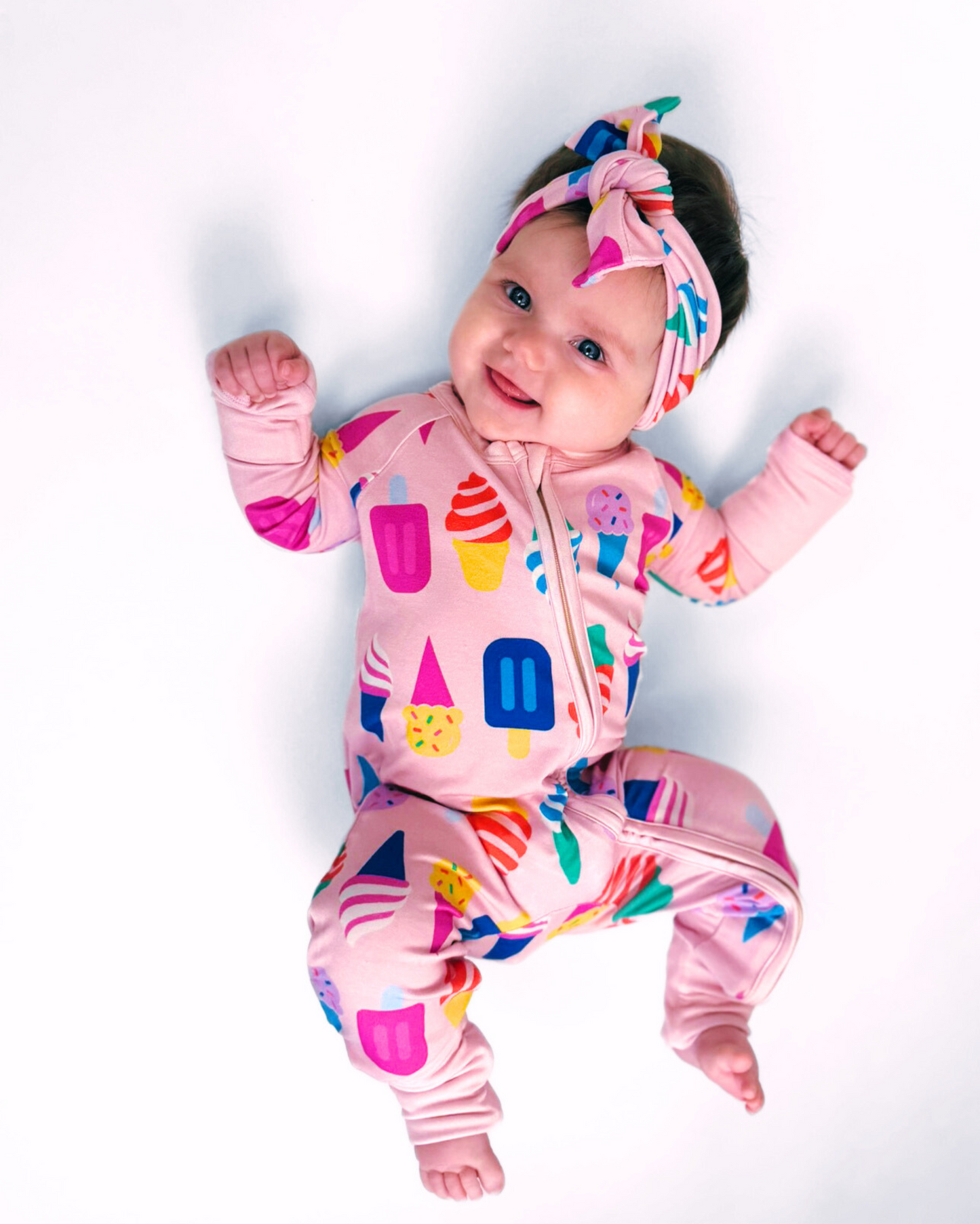 Cute baby in a pink onesie with colorful ice cream prints and matching headband, made from soft cotton.
