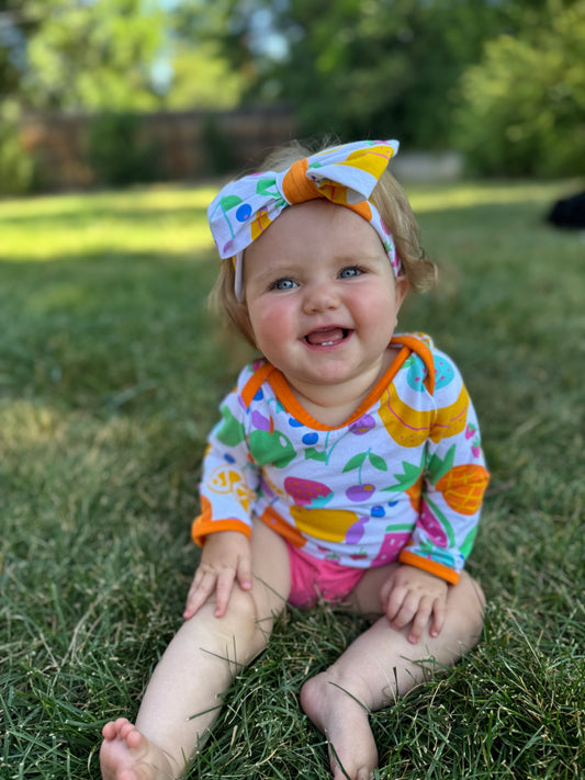 Toddler in colorful fruit-patterned long-sleeve shirt and pink shorts, sitting on grass with a cheerful expression.