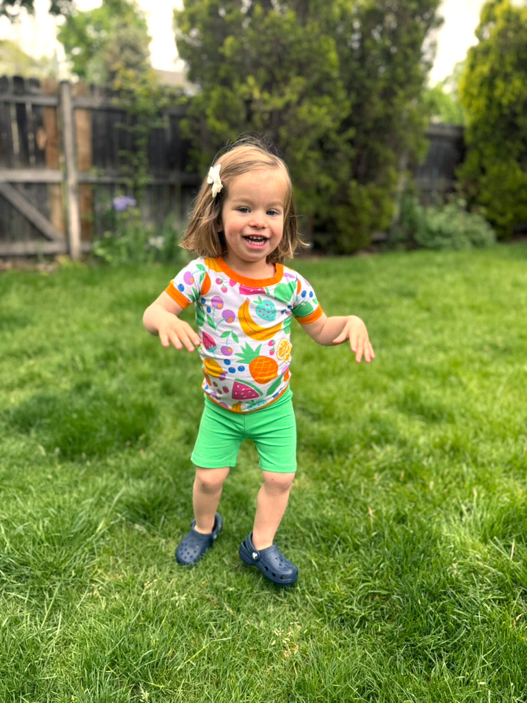 Toddler in colorful fruit-patterned shirt, green shorts, and navy crocs on grass, smiling happily.