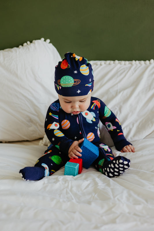 Baby wearing navy space-themed onesie and hat, playing with colorful building blocks on a bed.