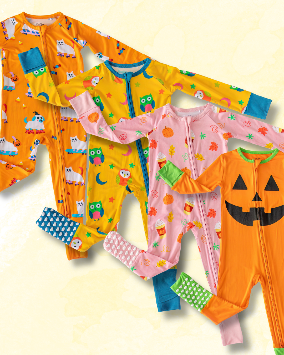Four children's Halloween-themed zip-up footed pajamas arranged on a light cream background. The pajamas feature orange, yellow, and pink colors with festive prints including jack-o'-lanterns, owls, pumpkins, ghosts, and candy corn.