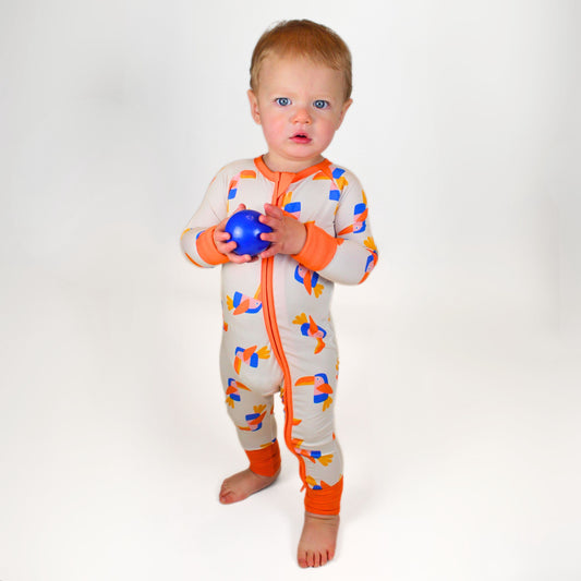 Baby wearing a colorful toucan-print onesie, featuring orange accents and a front zipper.