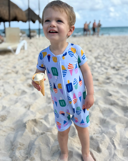 Toddler in a colorful ice cream-patterned onesie holding a cone on the beach.