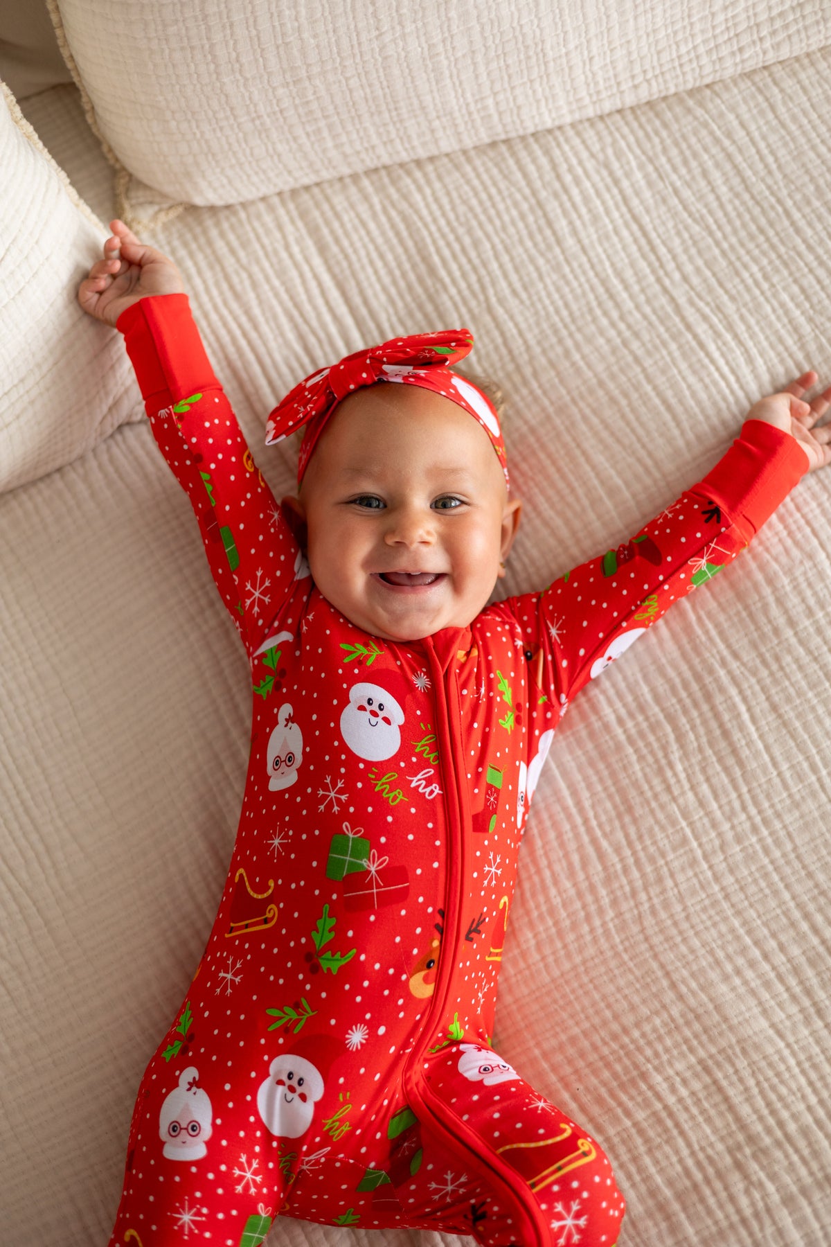 Adorable baby in a festive red Christmas onesie with snowmen and gift prints, featuring matching headband.