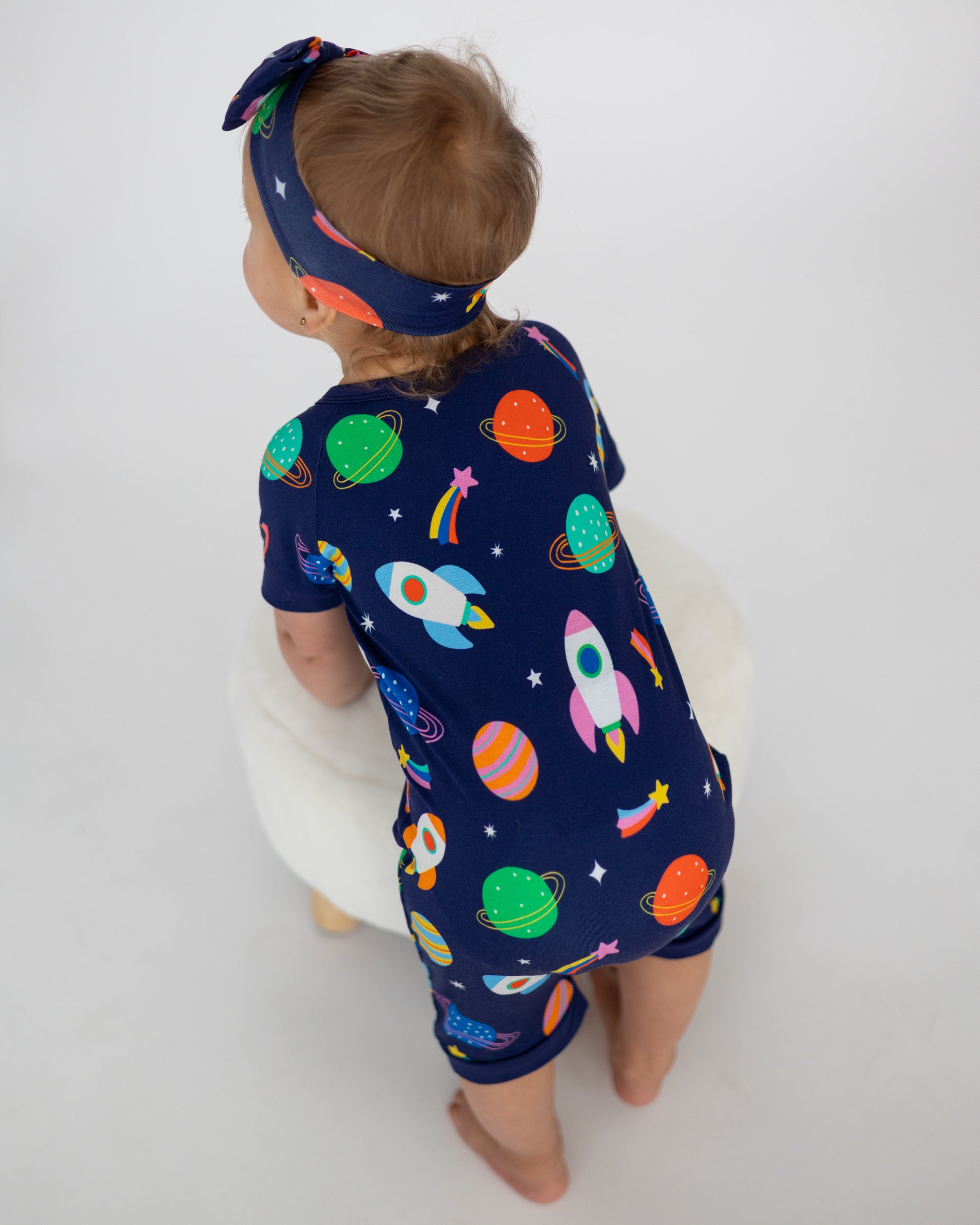 Toddler in a navy space-themed onesie with rockets and planets, paired with a matching headband.
