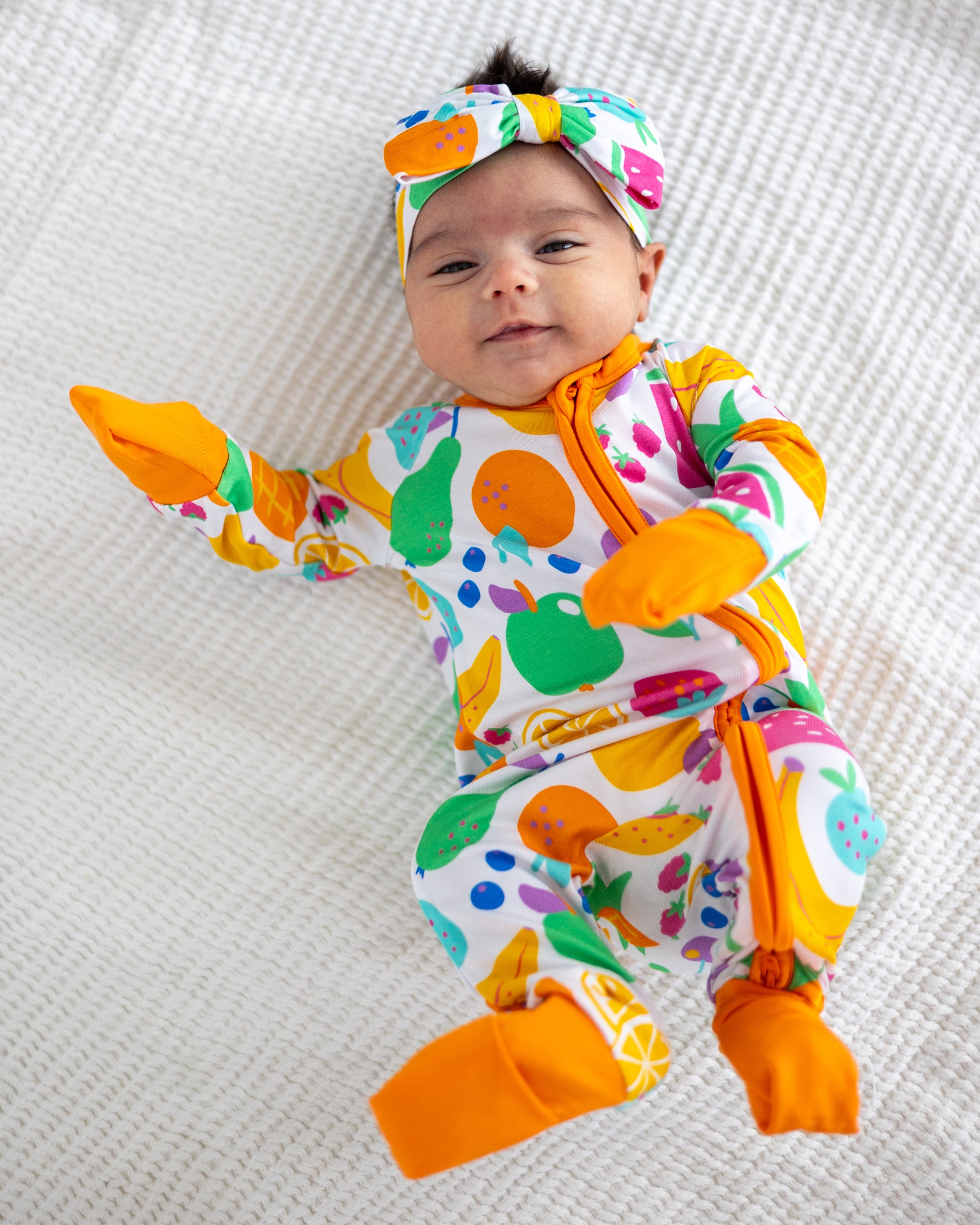 Colorful baby onesie with fruit patterns, orange accents, soft material, and matching headband.