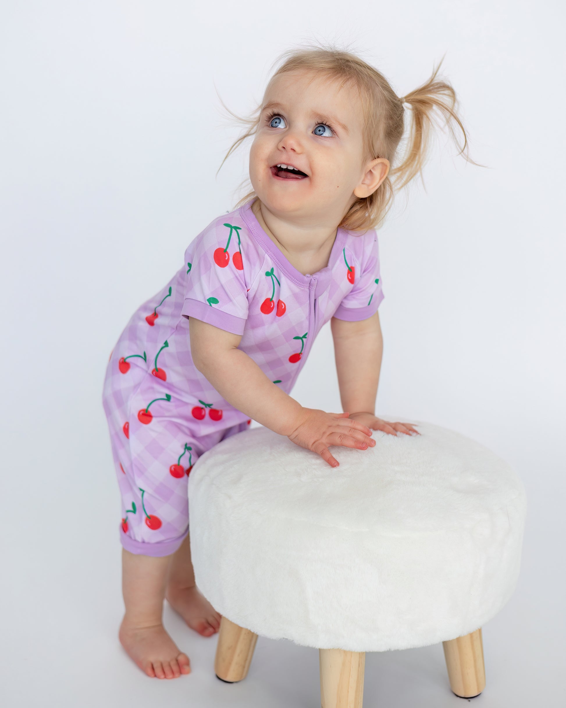 Toddler in purple cherry print romper, cozy and stylish, playing on a soft white stool.